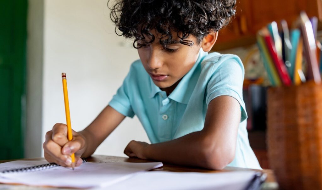 Boy with curly hair doing homework with pencil in notebook