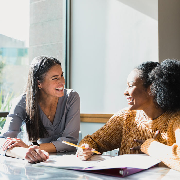 Two female students laughing while working on homework next to large sunny window