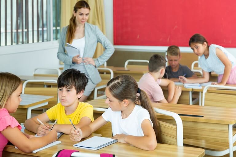 teacher stands and observes schoolchildren performing group tasks in classroom